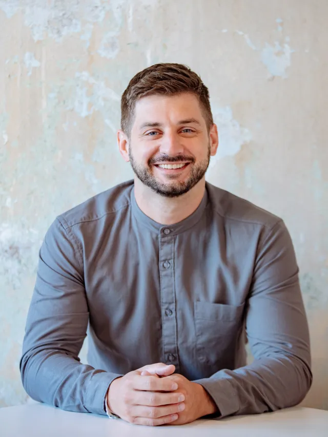 Alexander Stoffel, smiling, seated at a table in front of a pale, weathered concrete wall.