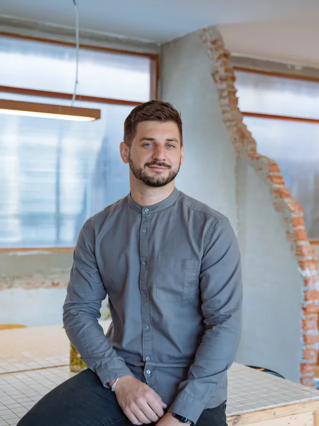 Alexander Stoffel seated on a workbench in a bright studio with exposed brickwork and tall windows.